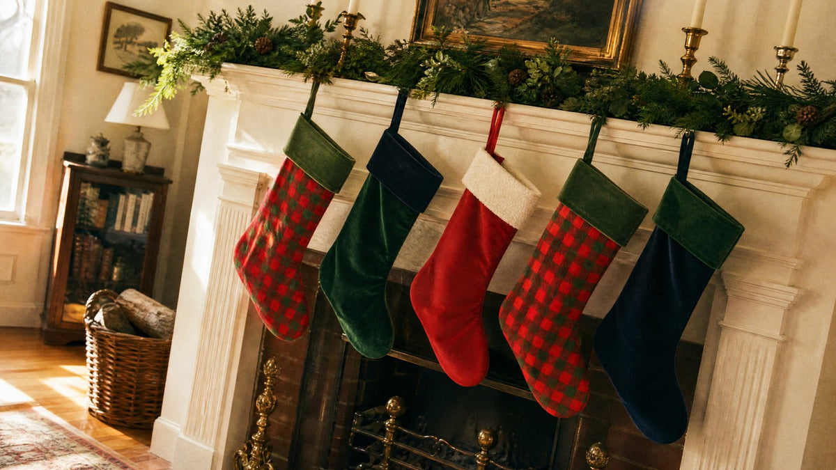 Five Christmas stockings in gingham check, green velvet and a white plush cuff hung in a row across a cream painted mantel with cedar garland