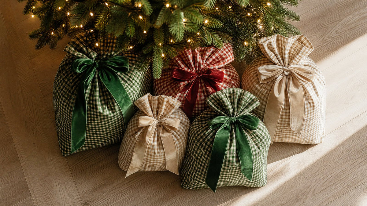 The base of a Christmas tree surrounded by five gingham check fabric gift sacks of different sizes, each tied with a velvet bow, instead of a tree skirt