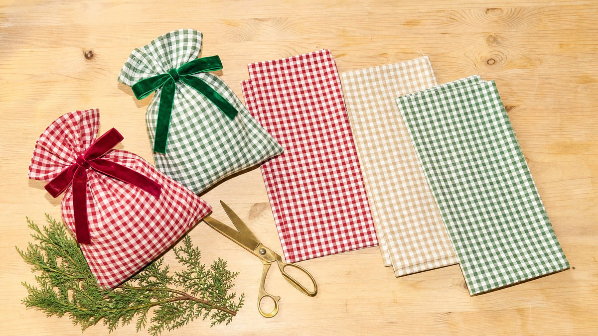 Five gingham check fabric gift sacks on an oak table, two filled and tied with velvet bows and three folded flat, with brass scissors and cedar