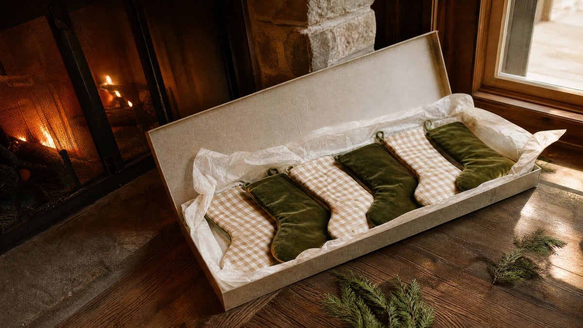 Six gingham and green velvet Christmas stockings laid in tissue paper inside an open fabric covered keepsake storage box on a wooden floor