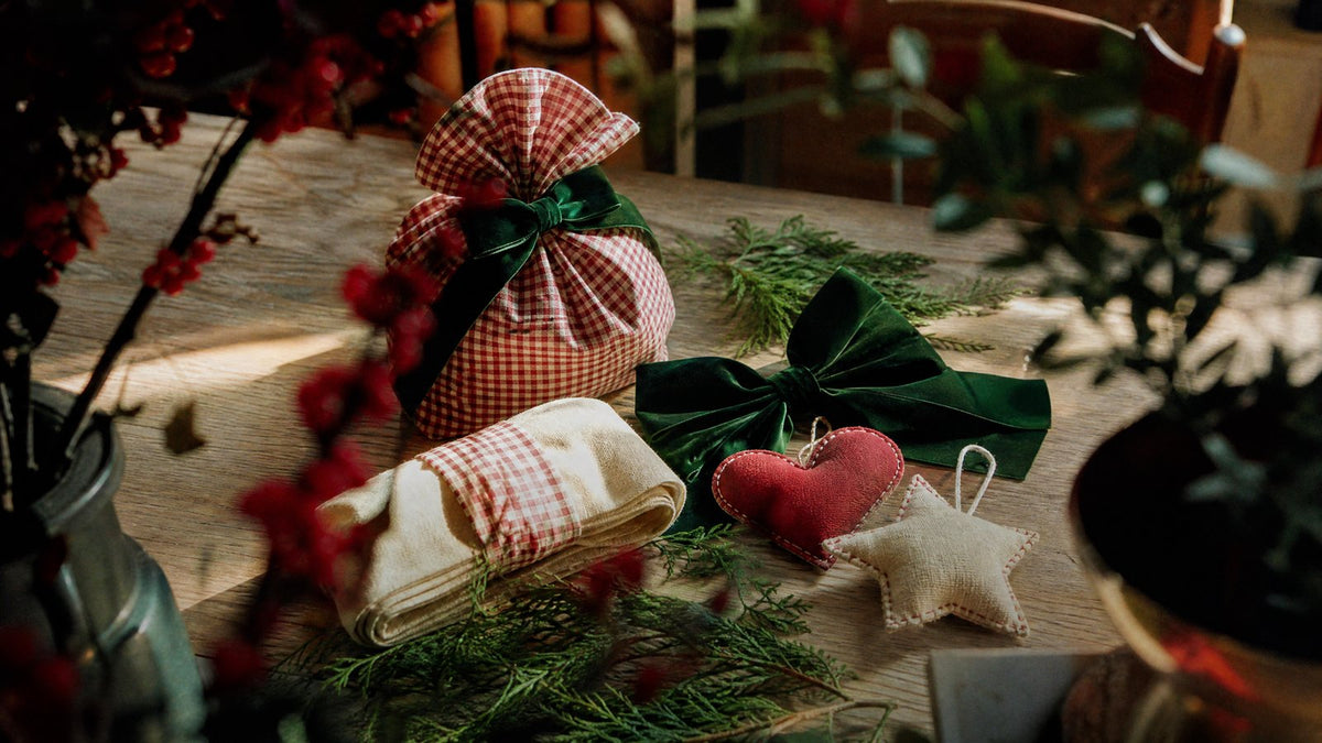 A selection of small Christmas gifts on an oak table: gingham fabric gift sacks, a green velvet bow and two soft fabric ornaments with cedar