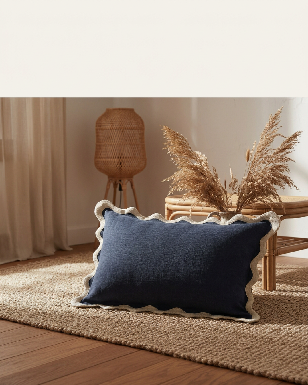 A navy linen cushion with a cream scalloped wavy trim on a jute rug with dried pampas behind